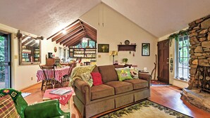 Una sala de estar acogedora con techo de madera, chimenea de piedra y zona de comedor con mantel a cuadros rojo y blanco.