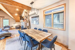 Un vaste espace dédié à la salle à manger, avec une grande table en bois et des chaises bleues, une cheminée en pierre et une grande fenêtre équipée de stores.