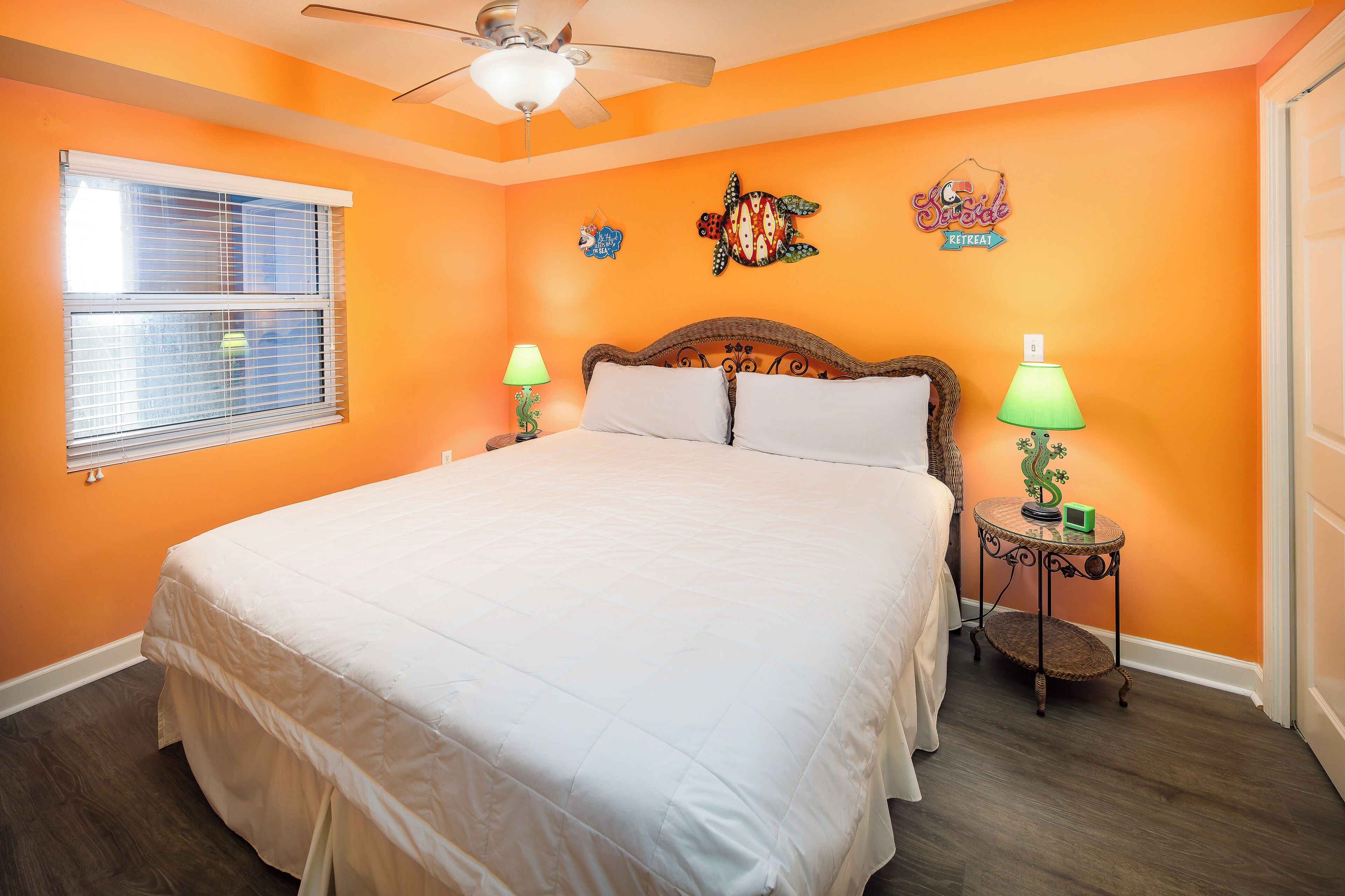 Un dormitorio con pared naranja, cama con ropa blanca, dos lámparas de noche, un ventilador de techo y una ventana con persianas.