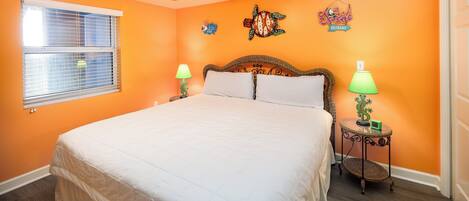 Un dormitorio con pared naranja, cama con ropa blanca, dos lámparas de noche, un ventilador de techo y una ventana con persianas.