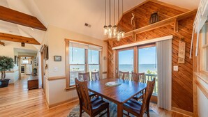 A spacious dining area with a wooden table and chairs, large windows with ocean views, and a wooden ceiling. - Doggone Perfect - Village at Nags Head, Soundfront, Golf Course View, Pool, Hot Tub, Pets OK (Nags Head)
