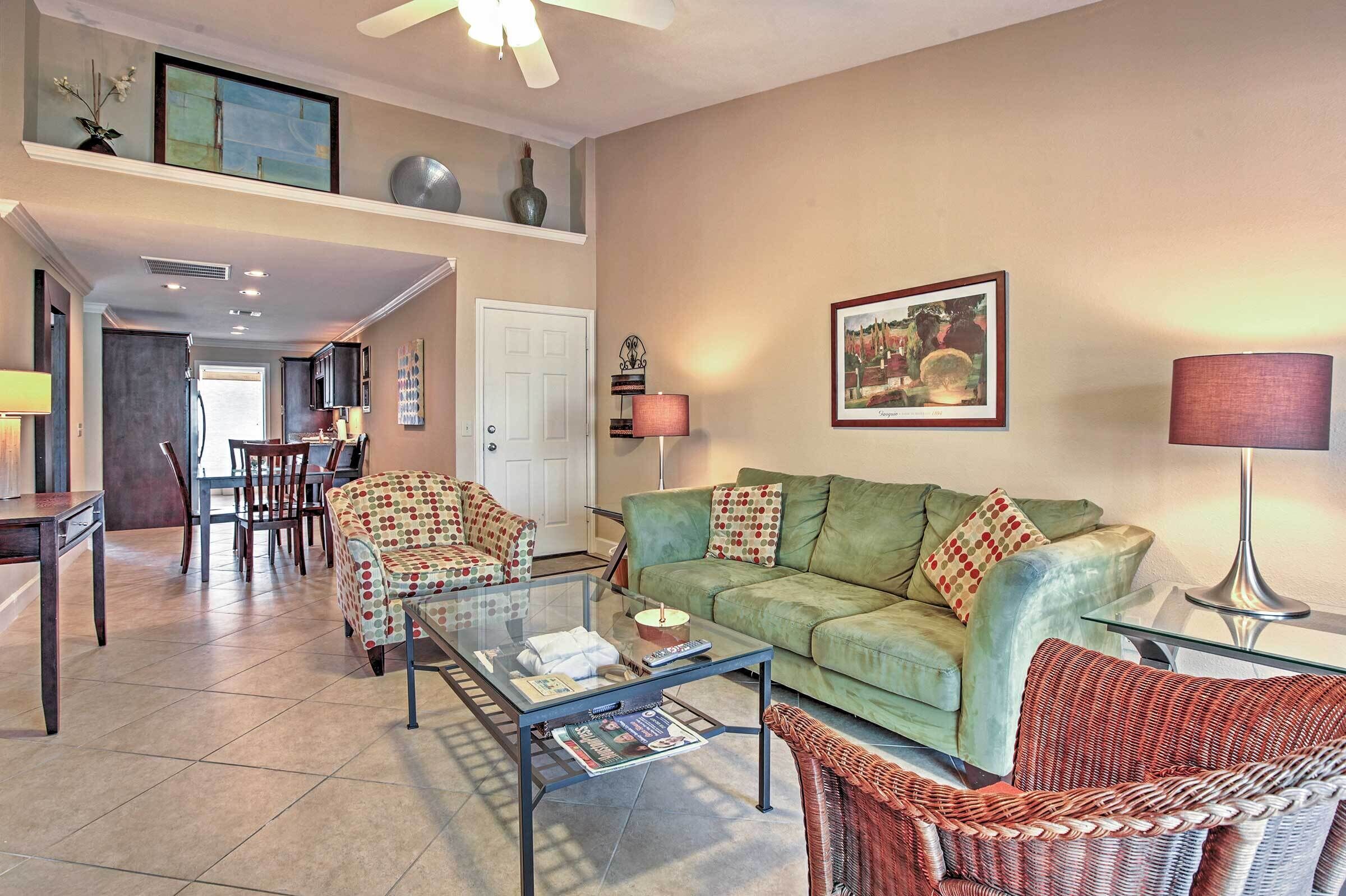 A spacious living room with a green sofa, a glass coffee table, and a wicker chair. There is a dining area with a table and chairs, and a kitchen in the background.