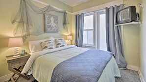 A bedroom with a bed, a nightstand, a television, and a window with curtains. - Steps to Avalon Bay: Sunny Catalina Island Home! (Avalon)