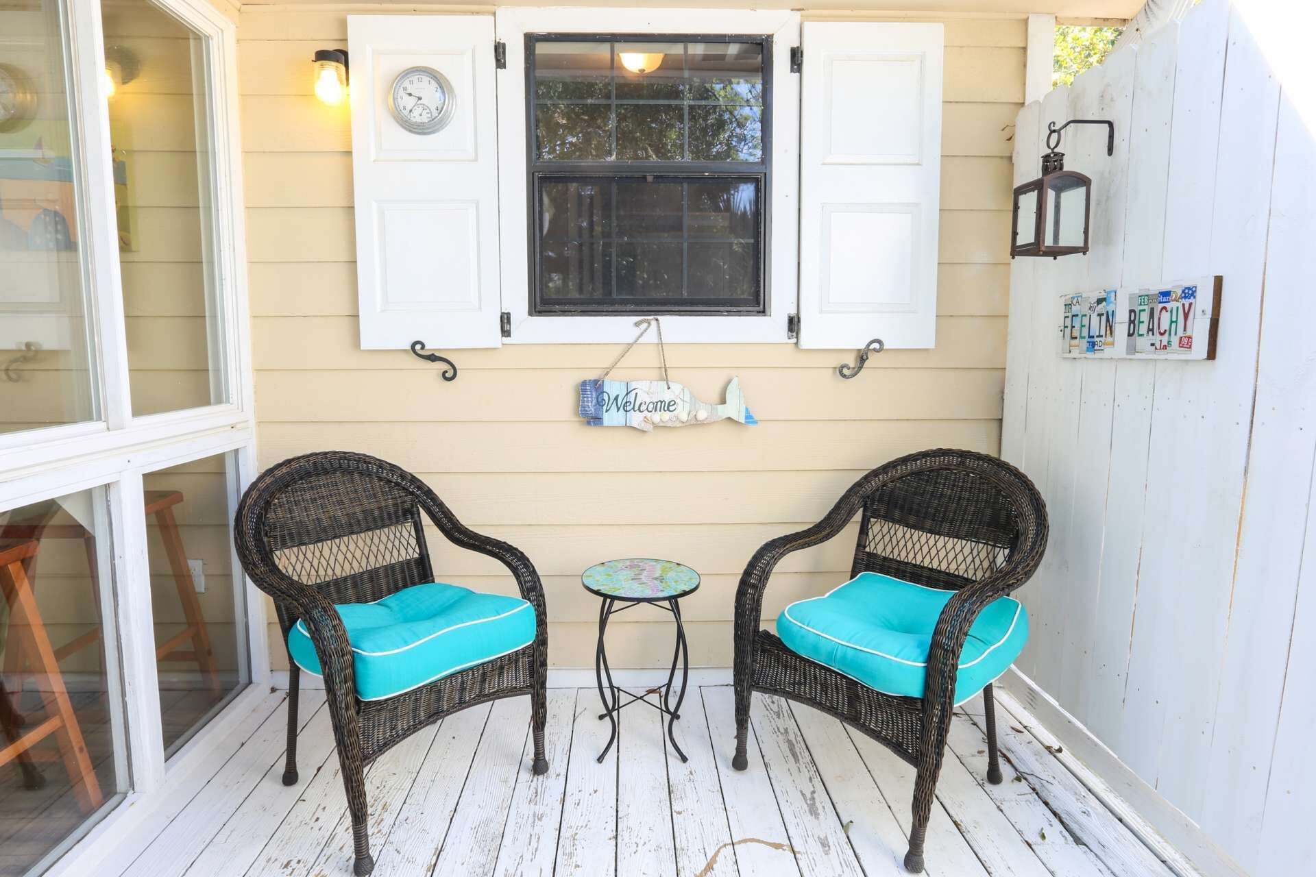 Een veranda met twee rieten stoelen met turquoise kussens, een kleine tafel en een raam met luiken.