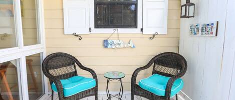 Een veranda met twee rieten stoelen met turquoise kussens, een kleine tafel en een raam met luiken.