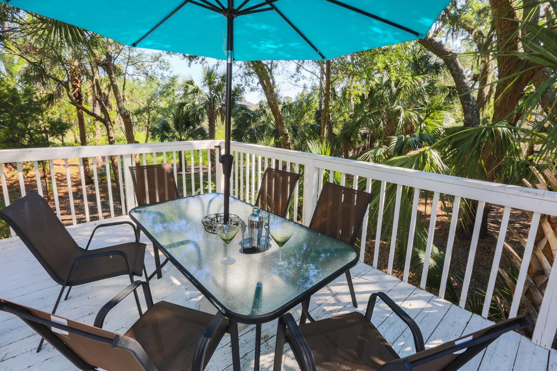 A glass-top table with four chairs, a blue umbrella, and a white railing on a deck surrounded by trees.