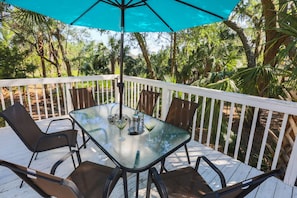 Mesa com tampo de vidro e quatro cadeiras, um guarda-sol azul e um corrimão branco em um deck rodeado por árvores.