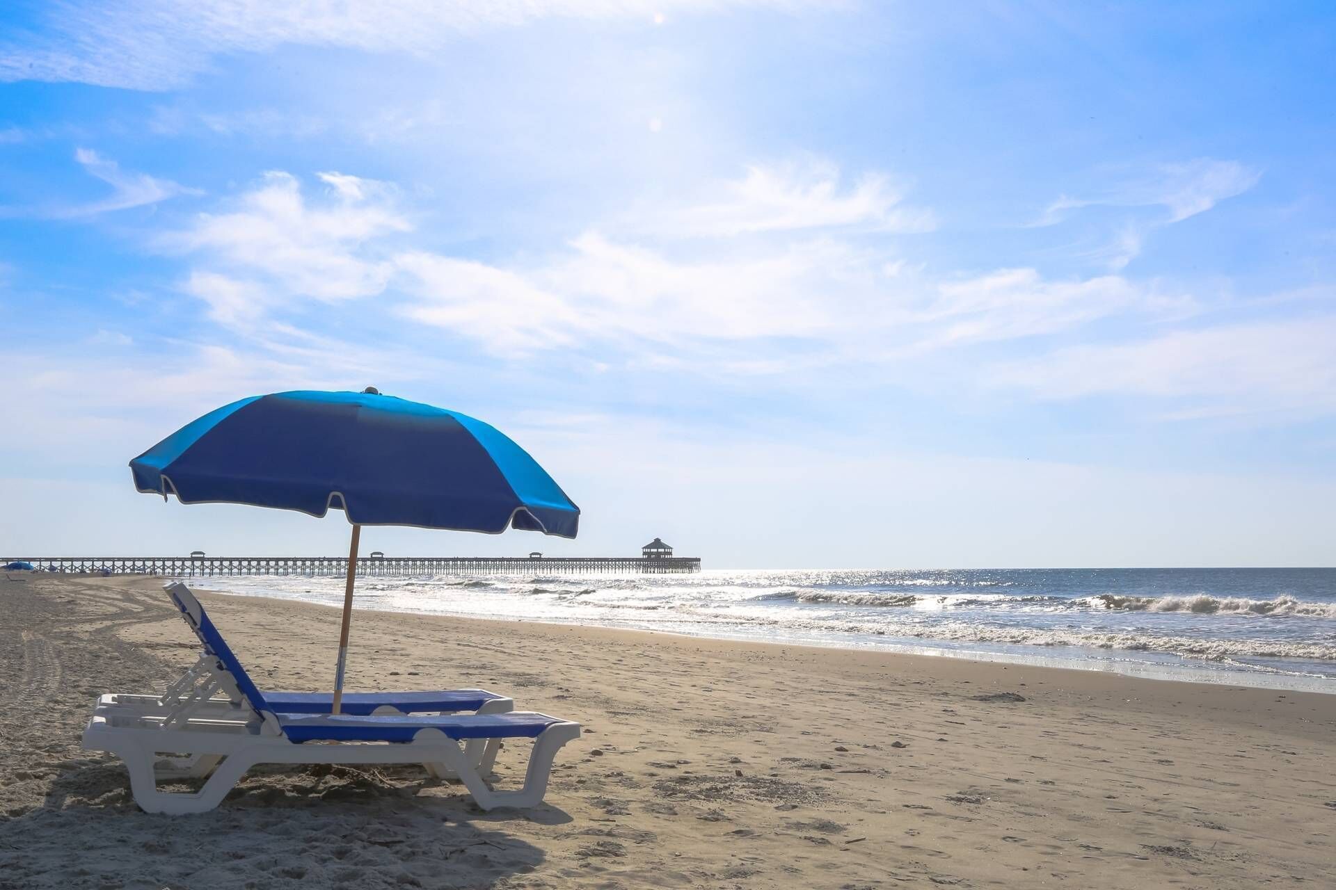A beach with a single lounge chair and a blue umbrella, a pier in the background, and the ocean.