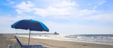 A beach with a single lounge chair and a blue umbrella, a pier in the background, and the ocean.