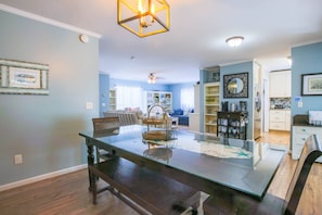 Uma área de refeições com mesa de vidro, cadeiras de madeira e um banco. Há prateleiras com objetos decorativos e uma cozinha ao fundo.