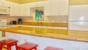 A kitchen with white cabinets, a granite countertop, a microwave, a toaster, a coffee maker, and a refrigerator. There are two red stools at the counter. - Cherry Grove Villas 204, Golf Villa Bliss near Ocean (North Myrtle Beach)