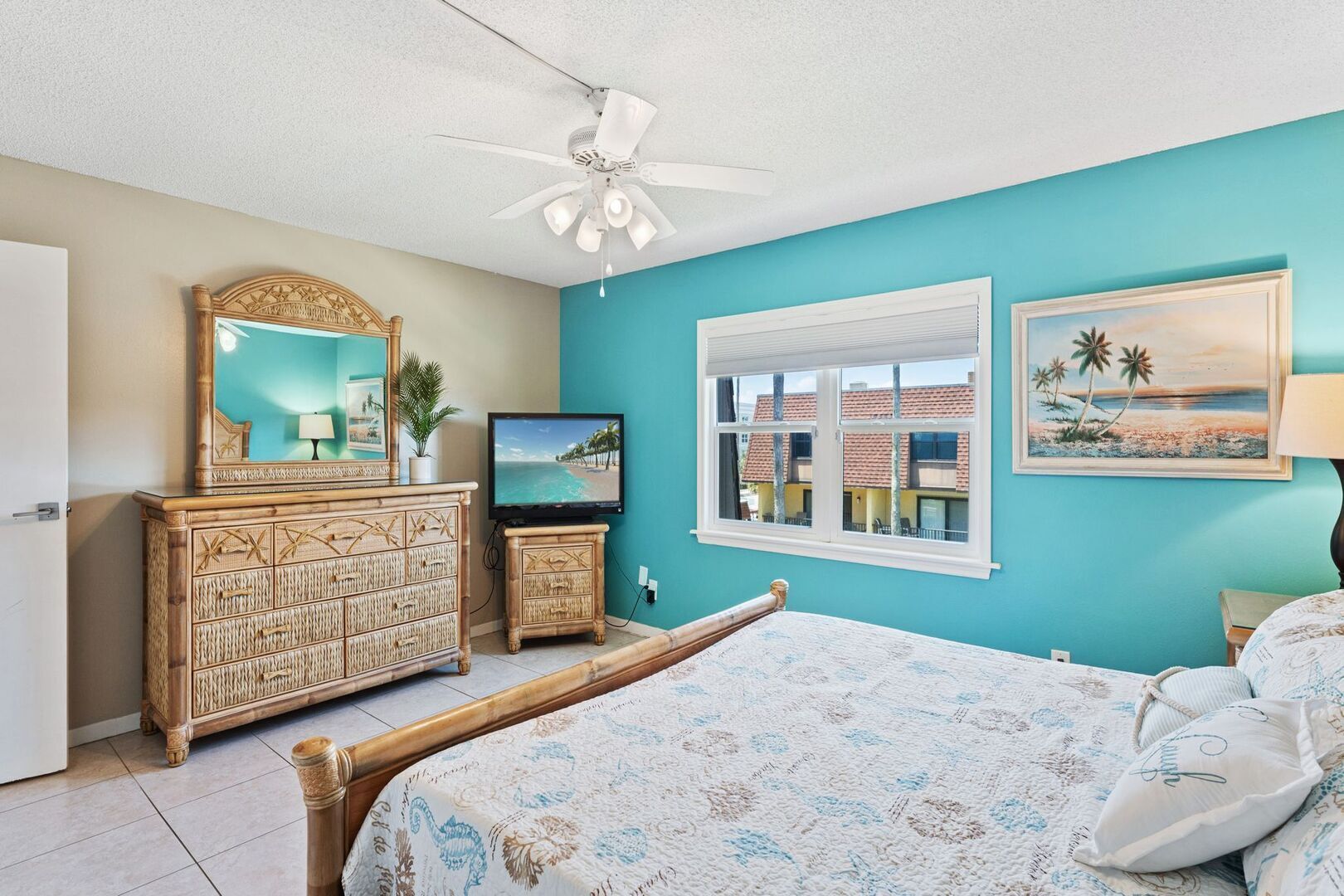 A bedroom with a bed, a wooden dresser, a television, a ceiling fan, and a window with a view of buildings.