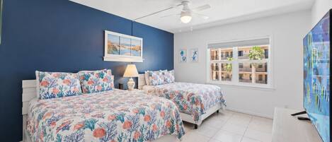 A bedroom with two beds, a ceiling fan, a window with a view of a building, and a TV on a stand.