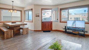 Un salón espacioso con suelo de madera, zona de comedor con banco, televisor sobre un mueble bajo y ventanas amplias con vistas al vecindario.