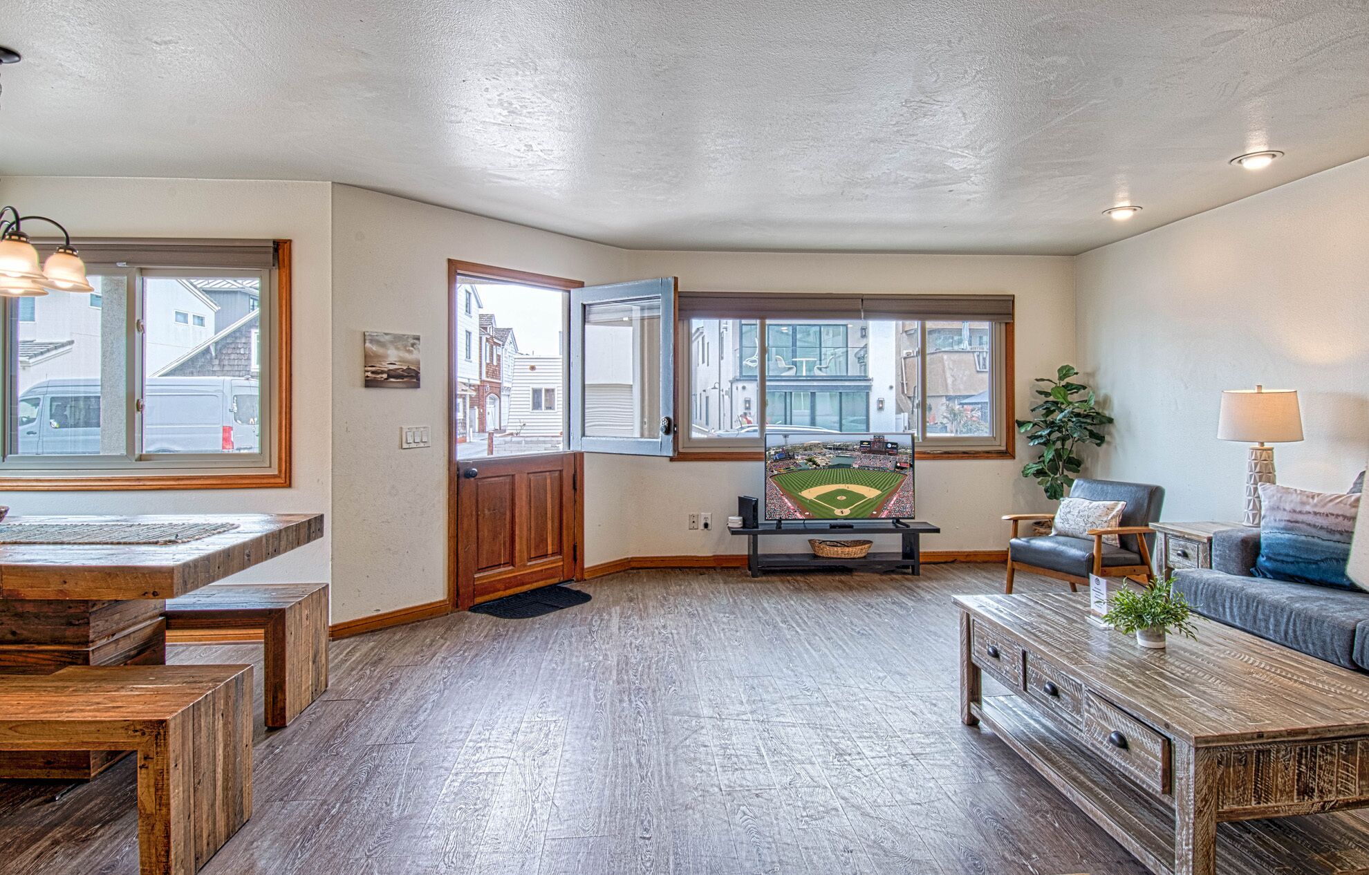 A spacious living room with a large window, a wooden coffee table, a sofa, and a television displaying a baseball game.