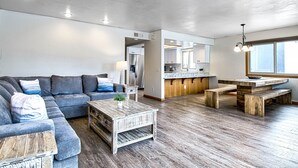Una sala de estar moderna con un sofá gris, una mesa de centro de madera y una cocina con armarios blancos y una mesa de comedor de madera.