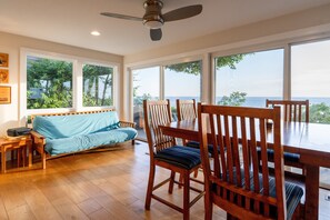 Ein großzügiger Wohnbereich mit einem Holzesstisch, Stühlen und einer Couch. Große Fenster bieten Blick aufs Meer.