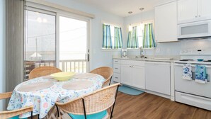 A compact kitchen with white cabinets, a microwave, and a stove. There is a dining table with a floral tablecloth and wicker chairs. - Comfortable sound-front duplex near Crystal Pier! (Wrightsville Beach)