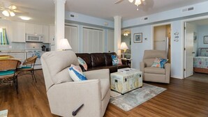 A living room with a beige sofa, a brown leather couch, and a small table with a vintage trunk. There is a ceiling fan, a microwave, and a dining area with wicker chairs. - Comfortable sound-front duplex near Crystal Pier! (Wrightsville Beach)
