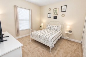 Une chambre à coucher avec un lit en métal blanc, une moquette beige, une fenêtre avec des rideaux et des cadres photo accrochés au mur.