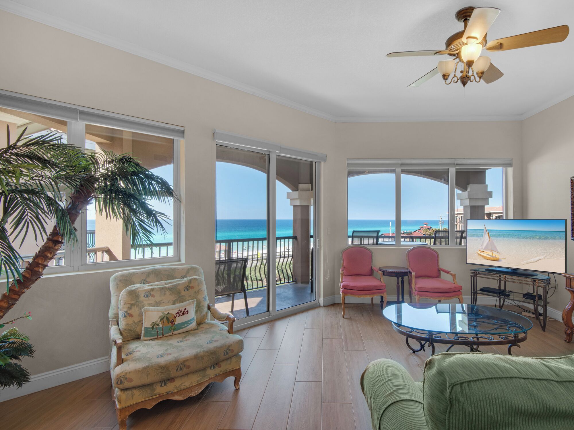 A spacious living room with a large window overlooking a beach, a ceiling fan, and a television displaying a beach scene.