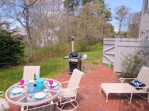 Une terrasse extérieure avec une table ronde en verre, des chaises en osier blanches et un barbecue.