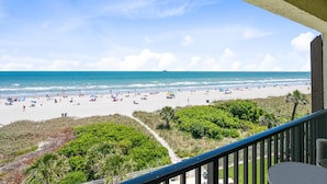 Une vue sur la plage depuis un balcon doté d’une balustrade, un sentier menant à la plage et quelques plantes au premier plan.