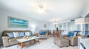 Un salon comprenant un canapé, un pouf et une table basse, un coin repas avec des chaises et une table, et une cuisine équipée d’un réfrigérateur et d’un four à micro-ondes.
