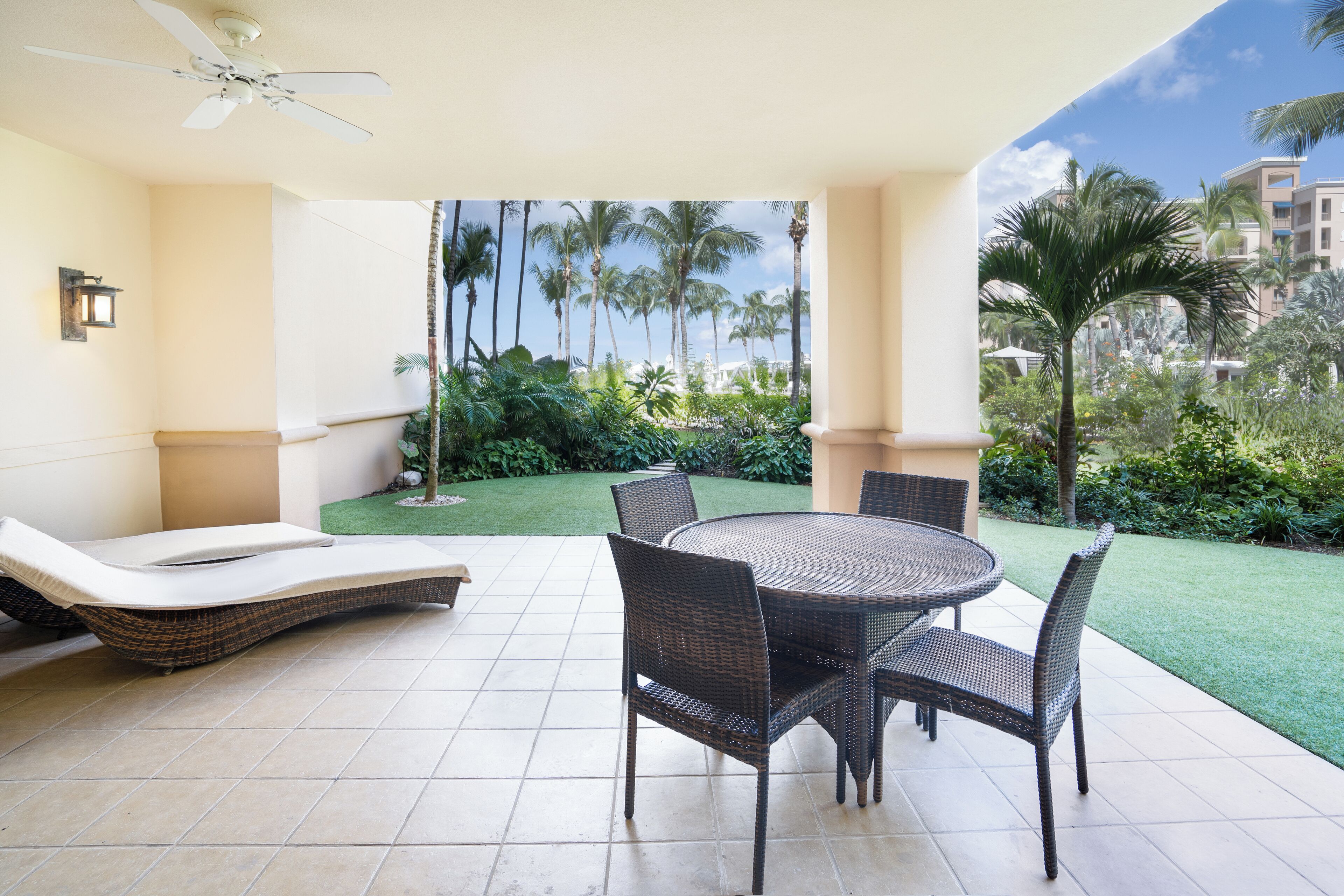 A covered patio with a round table and chairs, a lounge chair, and a view of a well-maintained garden with palm trees.