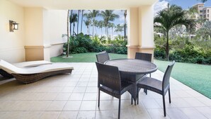 Un patio coperto con un tavolo rotondo e sedie, una poltrona da relax e vista su un giardino curato con palme.