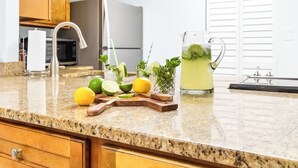 A kitchen counter with a granite countertop, wooden cabinets, a stainless steel faucet, and a glass pitcher of iced tea with mint leaves. - Fully Renovated Beachfront Penthouse with Breathtaking Ocean Views (Cocoa Beach)