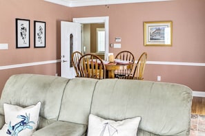 A living room with a beige sofa, two framed pictures, a dining table with chairs, and a door leading to another room.