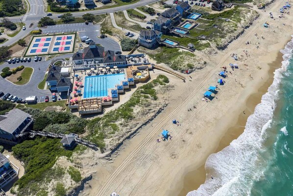 An aerial view of a coastal resort with a swimming pool, beach umbrellas, and a parking lot.