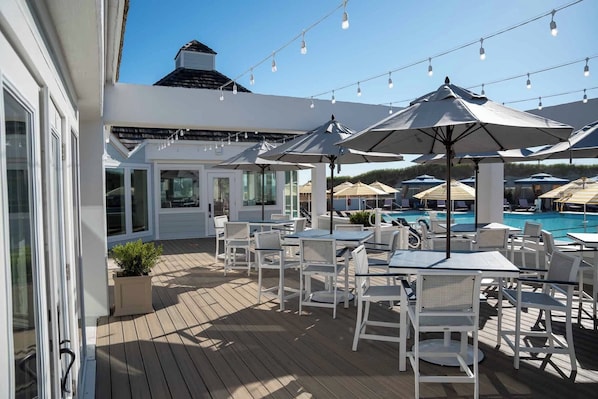 An outdoor patio with white tables and chairs, umbrellas, and a pool in the background.