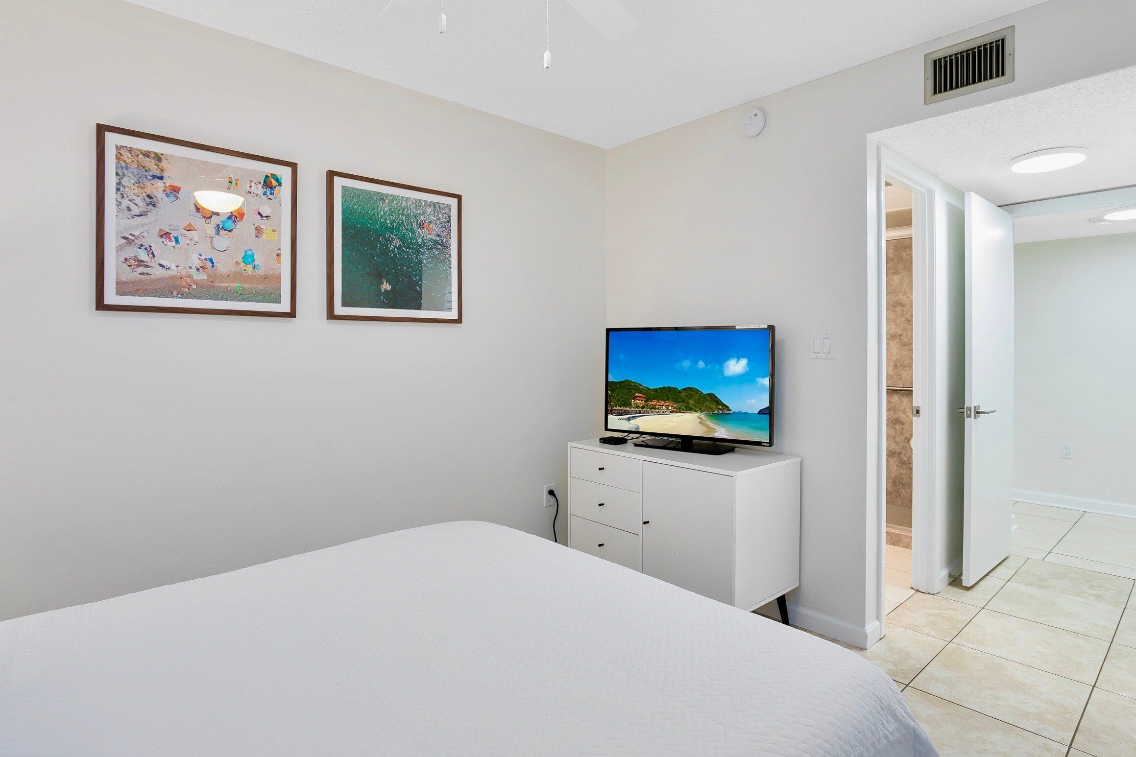 Un dormitorio con una cama, un televisor sobre un aparador blanco y dos cuadros en la pared.