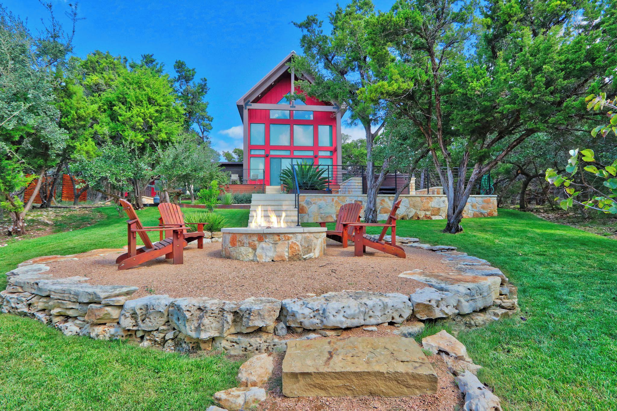A red wooden cabin with a stone fire pit and two wooden chairs in a landscaped garden.