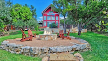 A red wooden cabin with a stone fire pit and two wooden chairs in a landscaped garden.