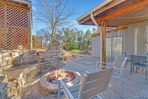 A stone fire pit with chairs, a wooden pergola, and a view of trees and a building.