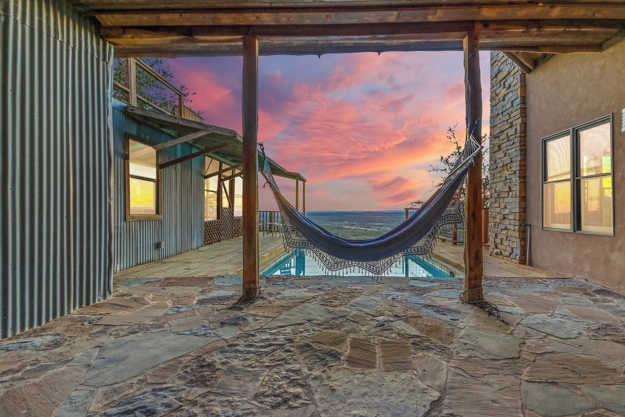 Una hamaca bajo una pérgola de madera con piso de piedra, con vista a una piscina y un paisaje con atardecer.