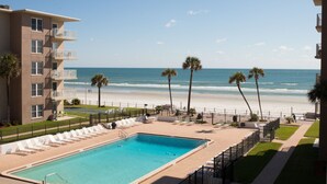 A pool area with a view of the beach, a building with balconies, and palm trees. - Surfs up in this Cute 2bed/2ba condo! Ocean view! No drive Beach in NSB (New Smyrna Beach)