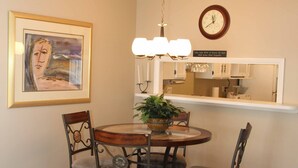 A dining area with a round table, four chairs, a chandelier, a clock, and a framed artwork on the wall. - Surfs up in this Cute 2bed/2ba condo! Ocean view! No drive Beach in NSB (New Smyrna Beach)