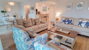 Ein modernes Wohnzimmer mit einem beigen Sofa, blauen Sesseln mit Muster und einem Couchtisch aus Holz, sowie einem Essbereich mit Korbmöbeln.
