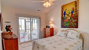 A bedroom with a bed, a nightstand, a lamp, a painting, and a sliding glass door leading to a balcony.