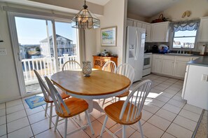 A kitchen with white cabinets, a wooden dining table, and chairs. There is a refrigerator, a window with a view of a house, and a door leading to a balcony.