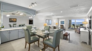 A spacious living area with a glass dining table, grey upholstered chairs, and a large window offering a view of the ocean.