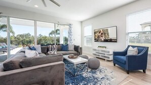 A modern living room with a large sofa, a coffee table, and a blue armchair. There is a TV mounted on the wall and a large window providing a view of the outdoors.