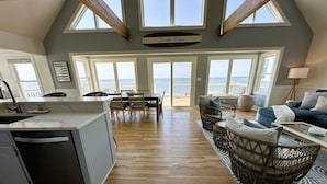Een ruime woonkamer met keuken, eethoek en zithoek bij een groot raam met uitzicht op de oceaan.