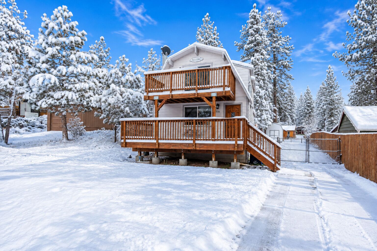 Une cabane à deux étages avec une terrasse en bois et une pancarte « Bienvenue », entourée d’arbres couverts de neige.