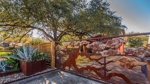 Ein schmiedeeisernes Tor mit rustikalem Design, das zu einem Garten mit unterschiedlichen Pflanzen und einer Holzkonstruktion im Hintergrund führt.
