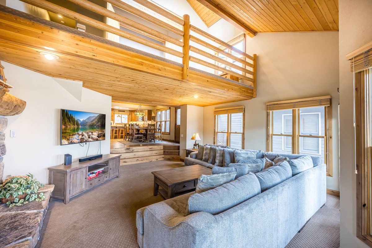 Ein geräumiges Wohnzimmer mit hohen Decken, Holzbalken, einem großen Sofa, einem Fernseher und einem Essbereich.